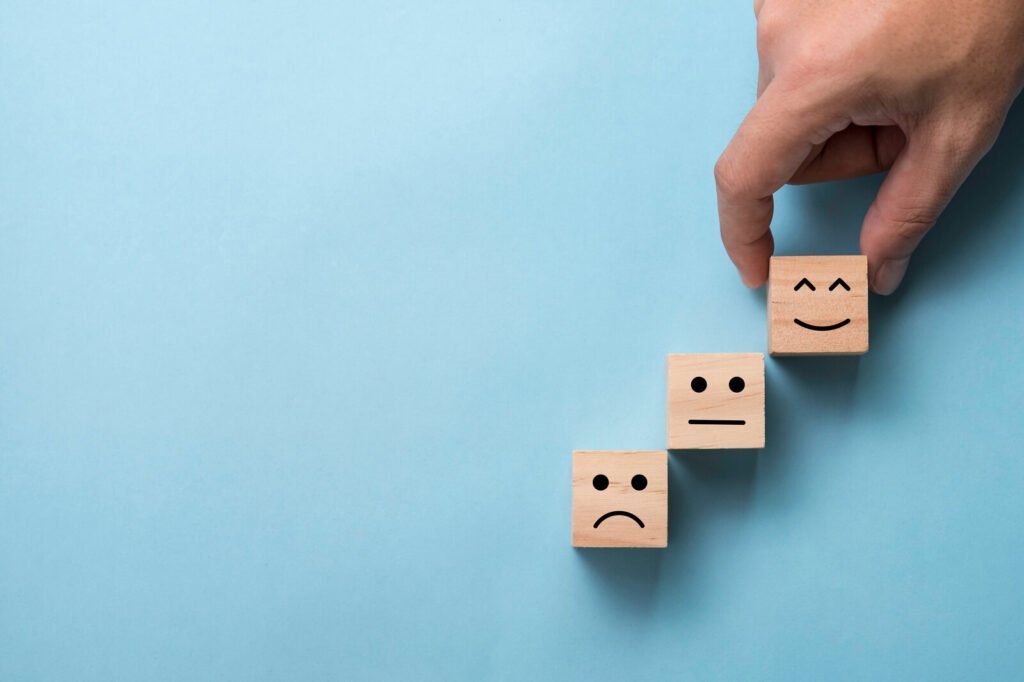 Wooden cubes arranged like a ladder. From bottom to top, each cube has the following emojis printed on its surface: sad, content, happy. A hand pinches the cube with a happy emoji.