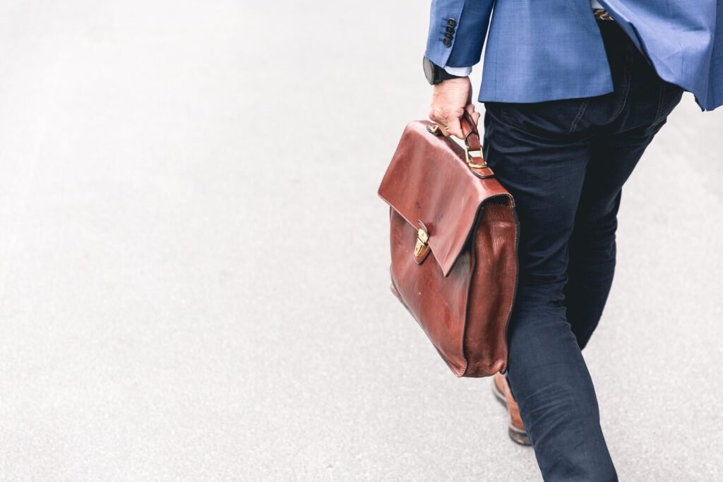 Below-waist shot of a walking man wearing dark jeans and a navy blue suit holding a leather briefcase